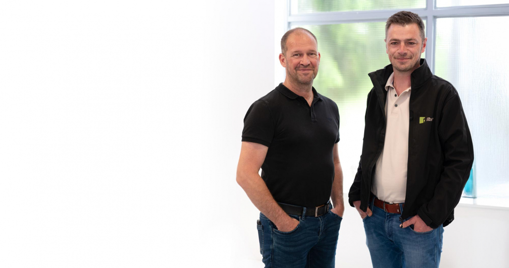 Auf dem Foto sind zwei Männer zu sehen - die Riedel Orthopädie Geschäftsführer. Sie stehen vor einem großen Fenster und lächeln leicht in die Kamera. Beide haben die Hände in den Hosentaschen.