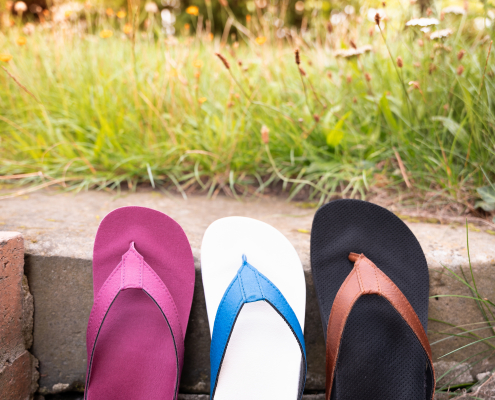 Auf dem Foto sieht man drei verschiedenfarbige Flip Flops, die auf einer Steintreppe angelehnt, an die zweite Stufe von oben stehen. Im Hintergrund sieht man Gras und den Ansatz von Büschen in der Unschärfe.