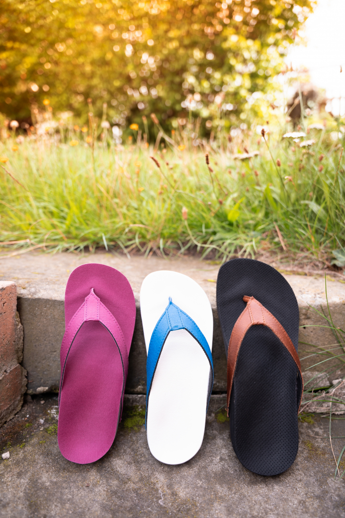 Auf dem Foto sieht man drei verschiedenfarbige Flip Flops, die auf einer Steintreppe angelehnt, an die zweite Stufe von oben stehen. Im Hintergrund sieht man Gras und den Ansatz von Büschen in der Unschärfe.