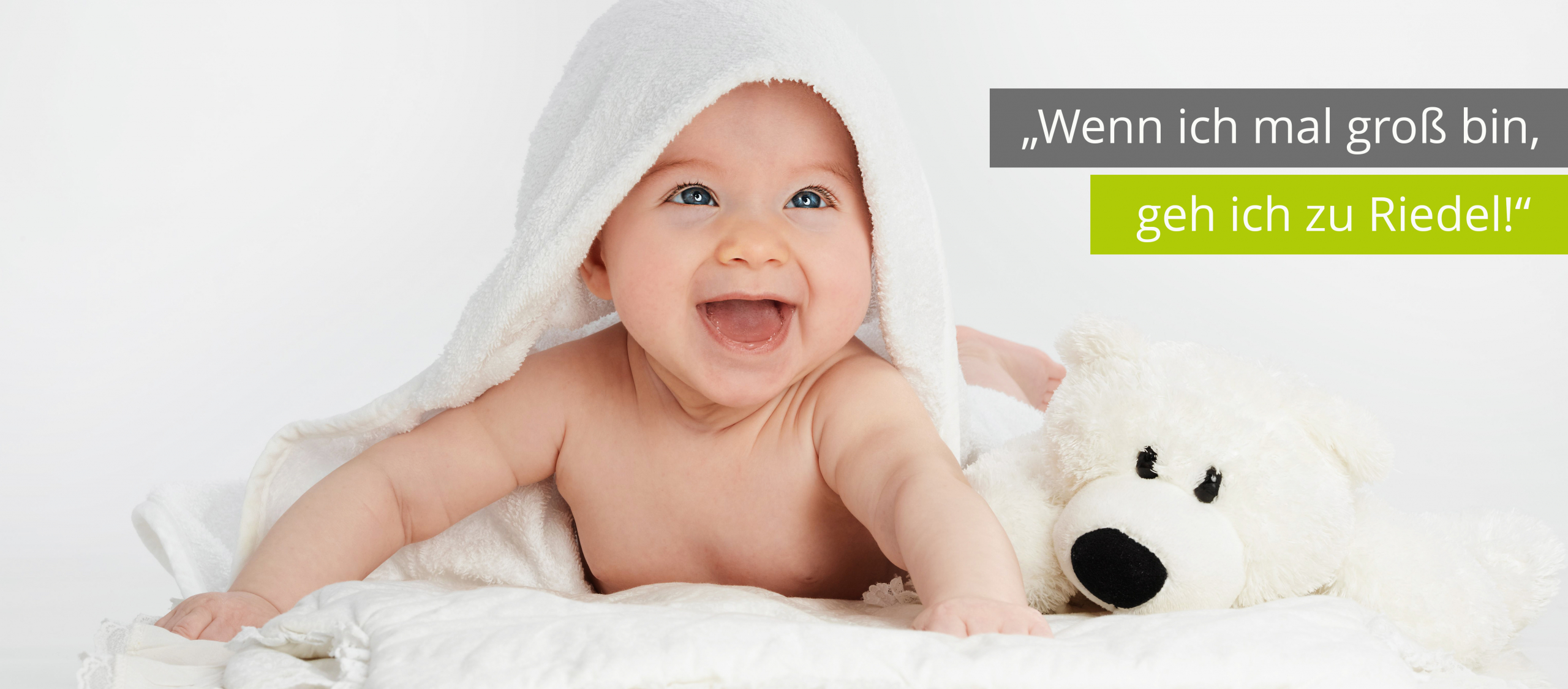 Das Bild zeigt ein Baby, welches lächeln auf einem Handtuch liegt und neben dem Baby liegt ein Kuscheltier. Darüber steht der Slogan: Wenn ich mal groß bin, geh ich zu Riedel.